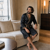 Modelo con bata negra para hombre con ribete dorado estampado, sentado en un salón elegante